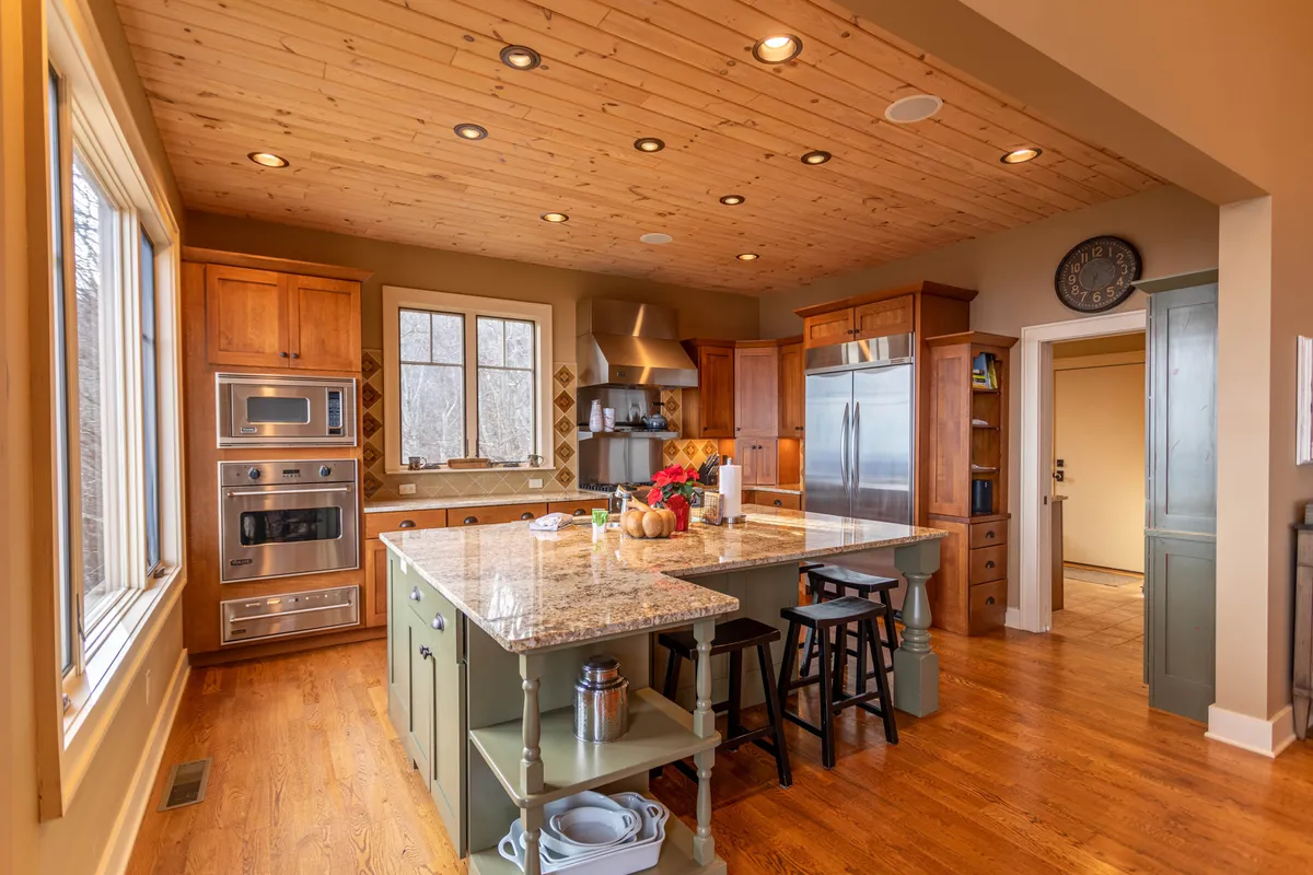A welcoming kitchen scene with a prominent granite countertop and large island, wooden cabinets lining the walls, and expansive windows providing natural light. The island offers ample space for meal prep and casual gatherings. The kitchen seamlessly connects to the surrounding living space, inviting communal activities.