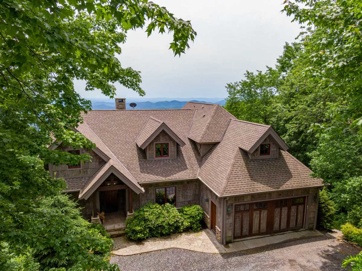The image showcases a stunning single-family mountain home nestled among lush greenery, with expansive layers of Blue Ridge Mountains stretching into the distance. The home features a spacious porch perfect for relaxing or gathering, with panoramic sunrise views and an inviting, tranquil atmosphere.