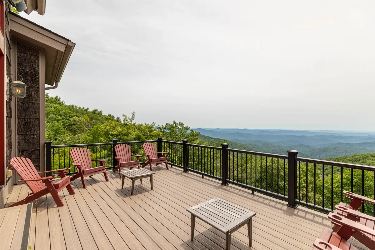 A wooden deck with red Adirondack chairs overlooks a stunning mountain view. The deck is equipped for relaxation and socializing, with a metal railing and clear sightlines to the surrounding landscape and sky.