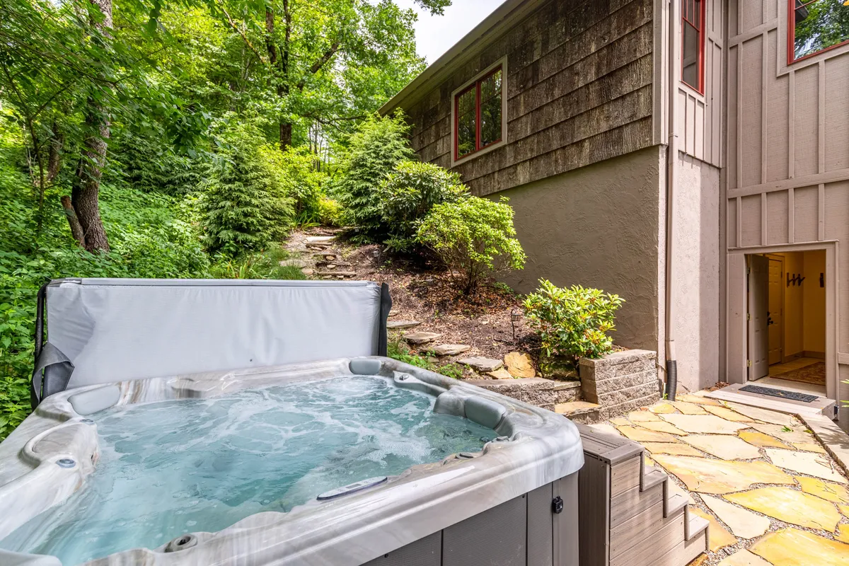 A luxurious hot tub set into a scenic deck area, bordered by a lush stone pathway and natural greenery, with the house nearby. The inviting setting is perfect for evening relaxation or social gatherings in the fresh mountain air.