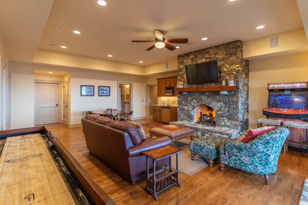 A cozy entertainment den featuring a stone fireplace, leather sofa, floral armchair, a large television, and a classic arcade game machine. The seating is arranged for comfortable socializing or relaxing while enjoying games or movies.
