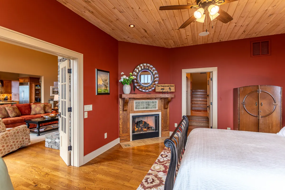 A warmly lit bedroom featuring a comfortable bed, a striking stone fireplace, and wooden ceiling beams. The room is designed for relaxation and intimacy, with a mirror and a cozy ambiance that makes it ideal for quiet nights or peaceful mornings.